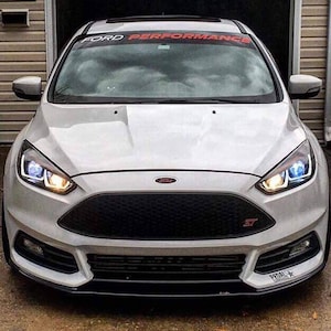 May include: A white Ford Focus ST with a black grille and red accents. The car has a black windshield banner that reads "FORD PERFORMANCE".