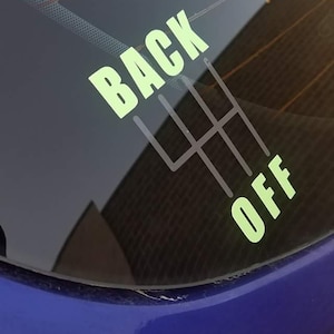 Puede incluir: Una pegatina verde y blanca en la ventana de un coche azul que dice "BACK OFF" con un patrón de palanca de cambios.