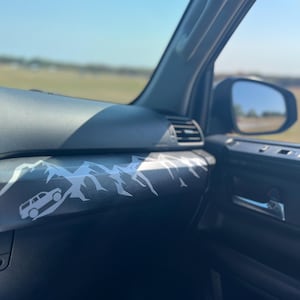 May include: Interior view of a vehicle's dashboard, featuring a black surface with a white mountain range and vehicle graphic decal. The dashboard includes air vents, a door handle, and a side mirror reflecting the outdoors.