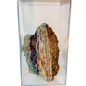 May include: A colorful, rough-hewn rock specimen encased in a clear acrylic display box. The rock features bands of brown, red, green, and white, creating a striking visual contrast. The display case provides a clean, modern presentation for the natural stone.