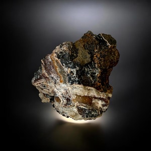 May include: A close-up of a rough, irregularly shaped rock formation. The rock displays a variety of colors, including brown, black, white, and hints of orange. The texture appears to be layered and somewhat crystalline, with a rough, uneven surface.