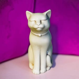 May include: A cream-colored cat figurine in a seated position with eyes closed in a serene expression. The cat features a smooth surface and wears a simple collar around its neck. The sculpture is rendered in a solid, light off-white tone.