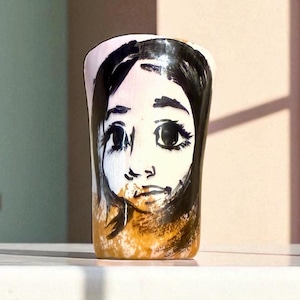 May include: A hand-painted ceramic tumbler featuring a stylized portrait of a woman's face. The artwork uses black and white brushstrokes for the facial features and hair, with a light pink and gold background.