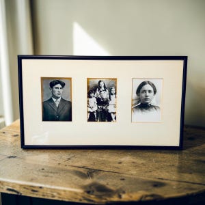 May include: A black rectangular picture frame with three vintage black and white photographs. The frame is on a wooden table. The photographs feature portraits of people, including a man in a suit and a group of children.