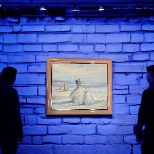 May include: An oil painting in a gold frame depicts figures on a beach. The artwork features a light color palette with figures in white dresses. The painting is displayed on a blue brick wall.