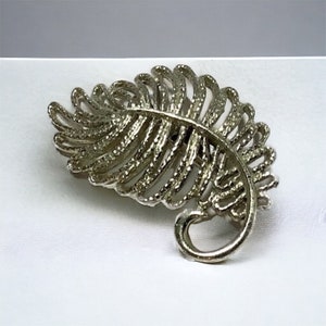 May include: A silver-toned brooch in the shape of a fern leaf. The leaf is detailed with a textured design.