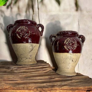 May include: Two antique-style ceramic vases. The upper portion is a deep burgundy color, while the lower half is a light beige. Each vase has two handles and a decorative emblem. The vases are displayed on a wooden surface.