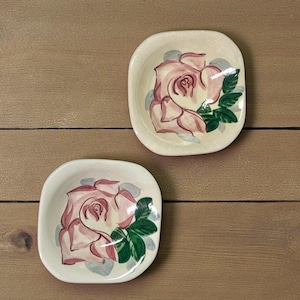 May include: Two small, square, off-white dishes with rounded corners. Each dish features a hand-painted pink rose with green leaves and gray accents. The dishes are set against a wooden background.