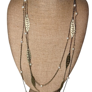 May include: A layered necklace with a bronze chain and leaf-shaped pendants. The necklace features small, white pearls interspersed along the chain. The leaf pendants have a detailed, cut-out design. The necklace is displayed on a tan mannequin.
