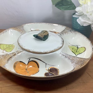 May include: A white ceramic serving dish with a gold rim and a removable center section. The dish has five sections, each decorated with a different leaf design in shades of green, orange, and brown. The dish is perfect for serving snacks, dips, or appetizers.