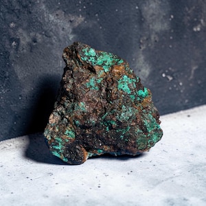 May include: A rough, irregularly shaped rock with a mix of brown, black, and turquoise-green colors. The surface has a textured appearance, with areas of the turquoise-green mineral contrasting against the darker, earthy tones.
