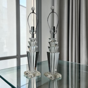 Op de afbeelding: Twee heldere kristallen tafellampen. Elke lamp heeft een geometrisch ontwerp met een gelaagde, facetgeslepen voet en een heldere kristallen finial. De lampen hebben een zilverkleurige metalen hals en een heldere glazen lamp.