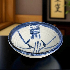 Puede incluir: Cuenco de cerámica hecho a mano con interior blanco y borde azul. El cuenco presenta un diseño abstracto azul en el centro. Probablemente se utiliza para servir alimentos o como pieza decorativa.