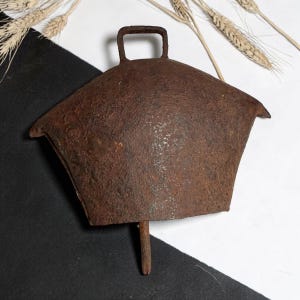 May include: A large, antique, rusty brown metal bell with a handle. The bell has a wide, flared shape and a clapper. The background is black and white with wheat stalks.