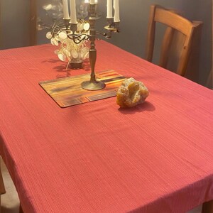May include: A coral-colored tablecloth covers a wooden table. A brass candelabra with lit candles sits on the table, along with a decorative wooden board and a large, light-colored rock. A wooden chair is visible in the background.