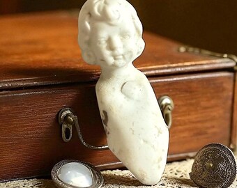 May include: A vintage porcelain doll head with a serene expression, featuring detailed facial features and curly hair. The doll head is off-white and rests near a wooden box and antique jewelry.