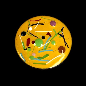 May include: A circular yellow button featuring a glossy finish with an abstract pattern of scattered dots and thin lines in various colors including black, white, red, green, blue, and purple, creating a vibrant and multi-colored surface design.