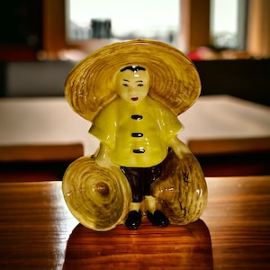 May include: A ceramic figurine of a person wearing a yellow shirt and a straw hat, holding two round straw baskets.