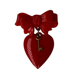 May include: A bright red brooch featuring a glossy bow at the top and a heart-shaped pendant suspended below. A small, antique-style brass-colored key is attached to the center of the red heart, creating a layered design with contrasting textures.