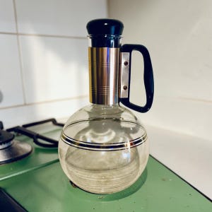 May include: A clear glass decanter with a round body and a black handle. The decanter has a black stopper and a gold-colored band around the neck. Two black and gold stripes encircle the body of the decanter.