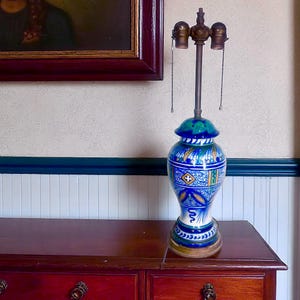 May include: A decorative ceramic table lamp with a blue, white, and yellow floral design. The lamp has a brass-colored stem and two light bulb sockets. The base is a wooden circle. The lamp is on a dark wood dresser.