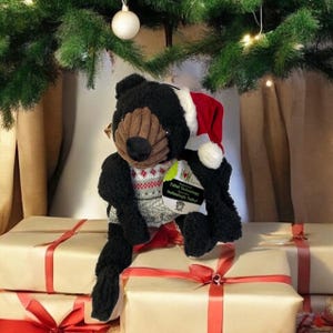 May include: A black teddy bear wearing a Santa hat and a sweater with a red and white pattern. The bear is sitting on top of wrapped gifts with red ribbons, under a Christmas tree. A tag on the bear reads "Taftut Technology".