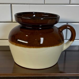 May include: A two-tone ceramic jar with a handle. The upper section is a glossy brown, and the lower half is off-white. The jar has a rounded body and a small, flared rim. The handle is a matching brown colour.