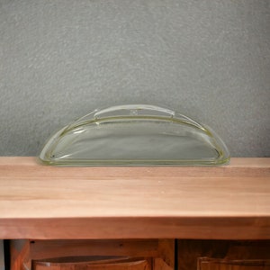 Op de afbeelding: Een transparante glazen koepelvormige deksel met een handvat. De deksel ligt op een houten oppervlak.