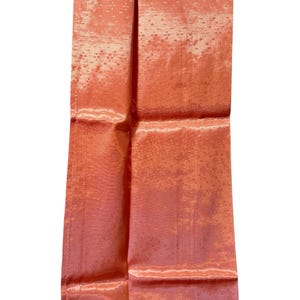 May include: A folded, rectangular piece of coral-colored fabric with a subtle, textured pattern. The fabric has a sheen, reflecting light and creating highlights. The item appears to be a decorative textile, possibly for home decor or fashion.