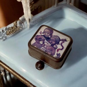 May include: A small wooden music box with a hand-painted image of three children playing music. The music box has a brown wooden exterior and a clear plastic lid. The image on the lid shows three children, two girls and a boy, playing musical instruments. The girl on the left is playing a guitar, the girl on the right is playing a violin, and the boy in the middle is playing a flute. The music box has a small wooden knob on the side that can be turned to wind the music box.
