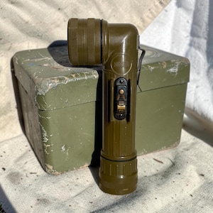 Puede incluir: Una linterna en ángulo recto de color verde oliva con un agarre texturizado y un interruptor negro. La linterna está delante de una caja verde a juego. La linterna tiene un clip y una cabeza acanalada.