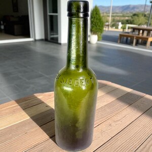 Könnte beinhalten: Eine antike grüne Glasflasche mit einem dunkelgrünen Verschluss. Das Wort "SIEGERT" ist auf der Flasche eingeprägt. Die Flasche steht auf einer Holzoberfläche.