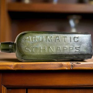 May include: A vintage green glass bottle, rectangular in shape, with the embossed words "AROMATIC SCHNAPPS". The bottle has a long neck and a flat body. It is displayed on a wooden surface.