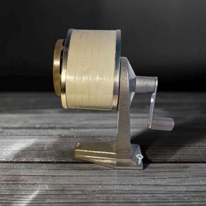 May include: A manual pencil sharpener featuring a cylindrical beige body with a metallic gold-toned front rim. The device is supported by a textured gray metal stand and includes a side-mounted hand crank with a small gray handle for operation.