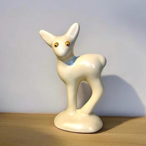 May include: A smooth, cream-colored ceramic figurine of a standing fawn. The deer has a glossy finish, pointed ears, and two distinct, round, amber-colored eyes that stand out against the pale body. It is positioned on an oval base.