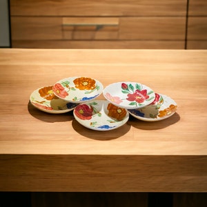 May include: A collection of six small, hand-painted ceramic plates. Each plate features a floral design with red, orange, and pink flowers, green leaves, and blue accents. The plates are arranged on a light-colored wooden surface.
