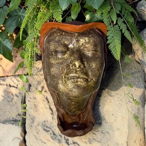 May include: A wall-mounted ceramic sculpture of a human face with closed eyes, featuring a metallic gold finish on the facial features and a contrasting matte terracotta-colored border and base. The sculpture has a hollow basin at the bottom.