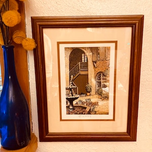 May include: A framed print of a courtyard scene with a fountain, stairs, and potted plants. The print is in a brown wooden frame with a white mat. The artist's signature is visible in the bottom right corner.