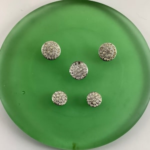 May include: Five silver-toned round buttons with clear rhinestones on a green background.