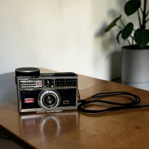 May include: A white ceramic mug with a black handle and the text "But First, Coffee" in a bold, black font. The mug is set against a blurred background, suggesting a cozy setting. The mug appears to be approximately 4 inches tall.