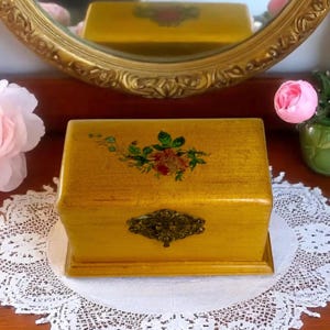 Puede incluir: Una caja de joyería rectangular de madera amarilla con un diseño floral en la tapa. La caja presenta un cierre de metal decorativo y se encuentra sobre un tapete de encaje blanco. Un espejo con marco dorado en el fondo.