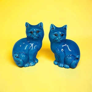 May include: Two matching glossy blue ceramic cat figurines in a seated position. Each cat features pointed ears, dark eyes, and a curled tail resting against its side. The smooth, reflective glaze covers the entire surface of both feline statues.