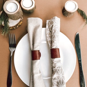 May include: Two white linen napkins with brown leather napkin rings on a white plate. The plate is on a brown surface with two wooden candle holders and a small sprig of greenery.