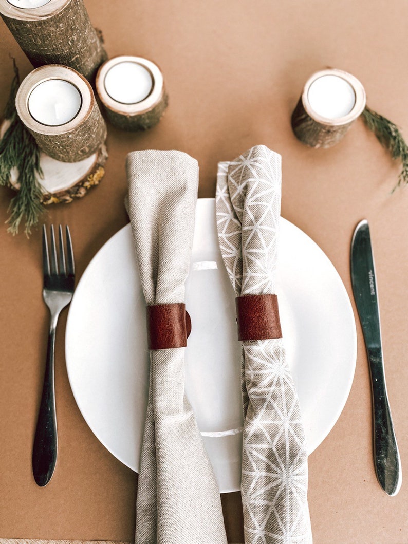 May include: Two white linen napkins with brown leather napkin rings on a white plate. The plate is on a brown surface with two wooden candle holders and a small sprig of greenery.