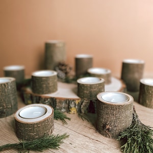 May include: A set of wooden candle holders with a rustic, natural look. The holders are made from tree bark and have a simple, elegant design. The candles are white and are placed in the center of the holders. The holders are arranged on a wooden surface with green sprigs of greenery.