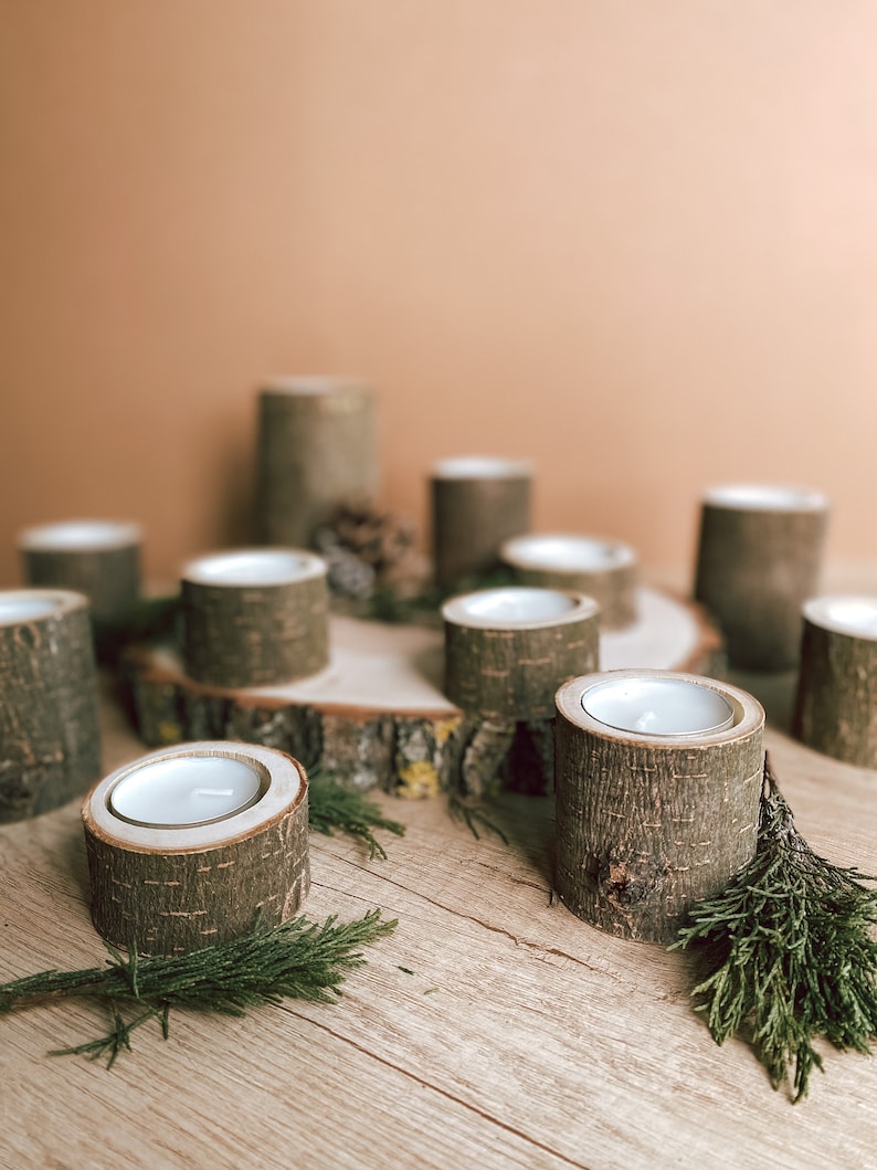 May include: A set of wooden candle holders with a rustic, natural look. The holders are made from tree bark and have a simple, elegant design. The candles are white and are placed in the center of the holders. The holders are arranged on a wooden surface with green sprigs of greenery.