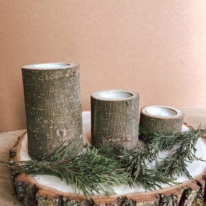 May include: Three wooden candle holders with a natural bark finish, arranged on a wooden slice with green foliage.
