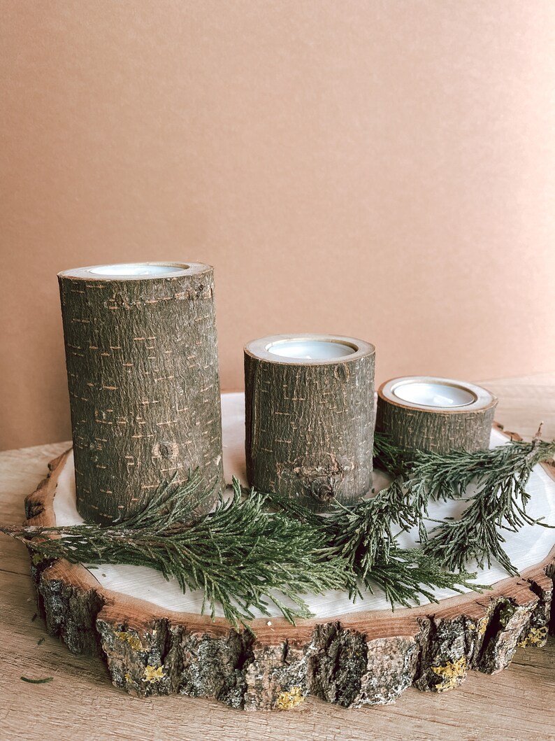 May include: Three wooden candle holders with a natural bark finish, arranged on a wooden slice with green foliage.