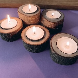 May include: Five wooden candle holders with lit tea lights. The candle holders are made of natural wood with a bark-like texture. The tea lights are white and are burning brightly.