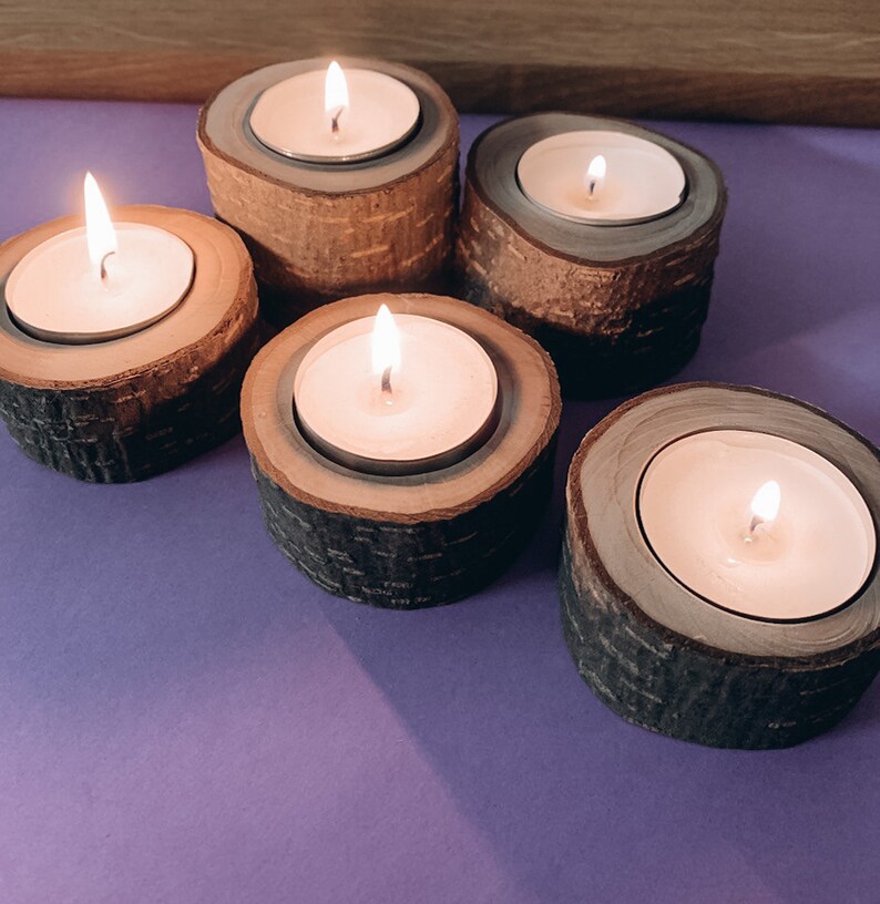 May include: Five wooden candle holders with lit tea lights. The candle holders are made of natural wood with a bark-like texture. The tea lights are white and are burning brightly.